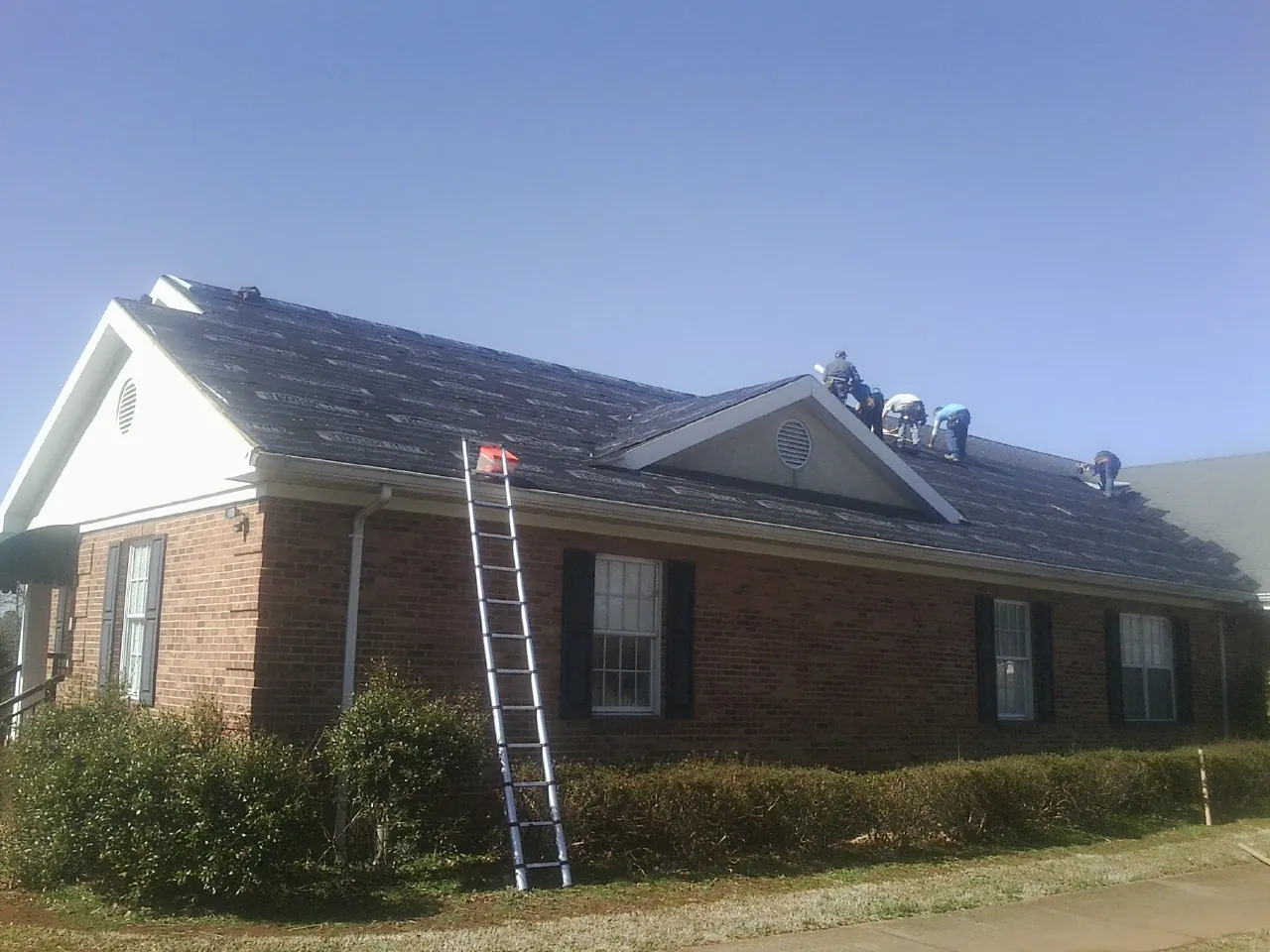 Crew tearing shingles off a brick ranch roof with a ladder set — Emergency Roof Repair work by our crew in Pinola