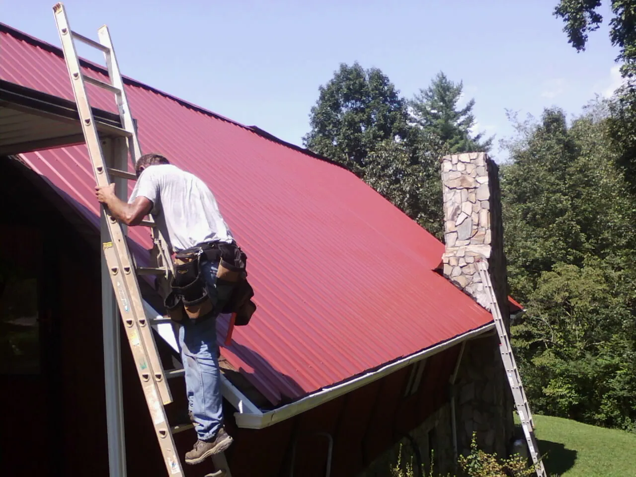 Roofer on a ladder beside a red metal roof and stone chimney — Soffit & Fascia Repair by Roofing Brothers in Pinola